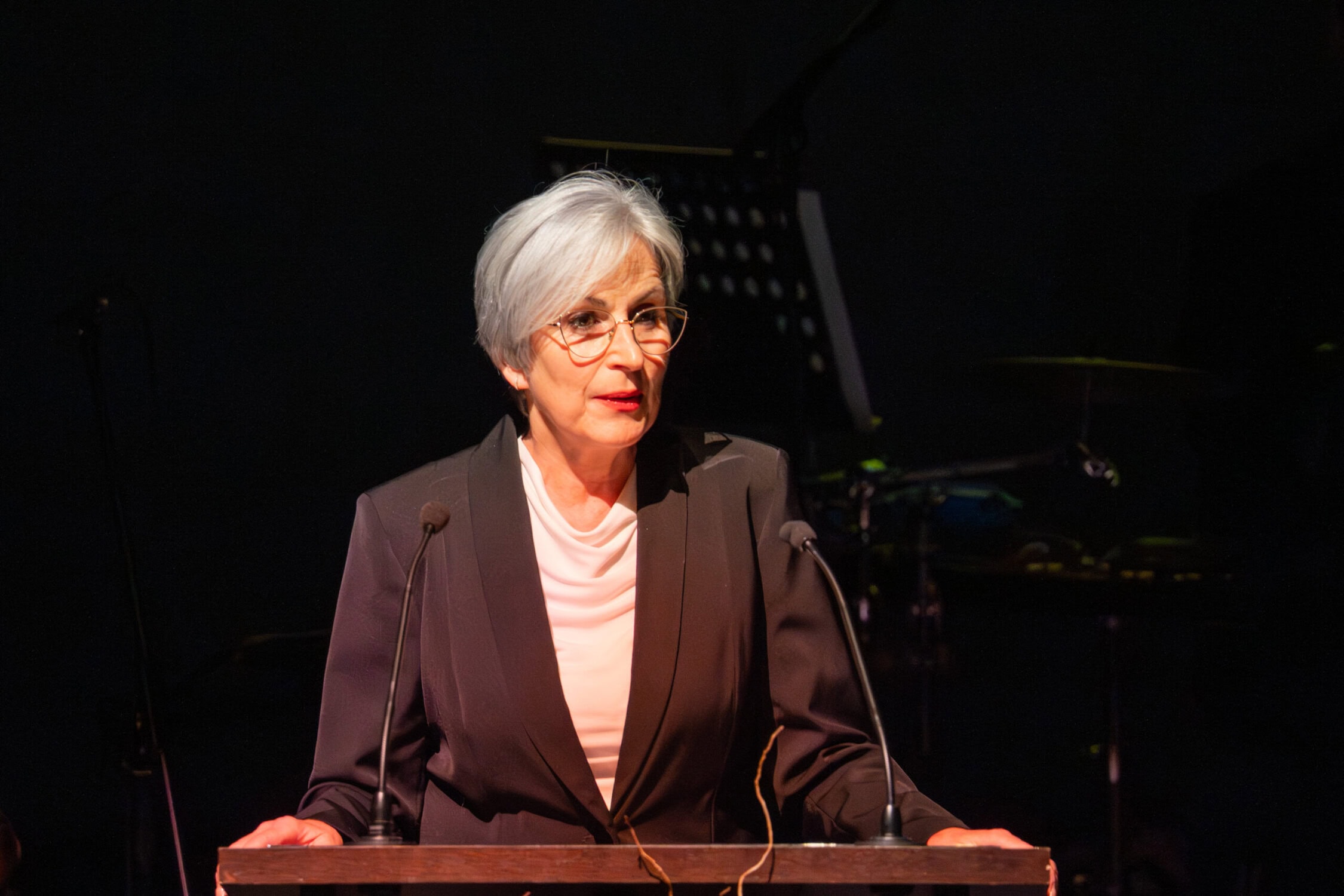 Richterin bei Rede auf Konferenz, Fokus auf Rechtsprechung und Justiz, professionelles Auftreten, formeller Anlass, dunkler Anzug, weiße Bluse, moderne Brille, im Hintergrund Klavier, Beleuchtung.