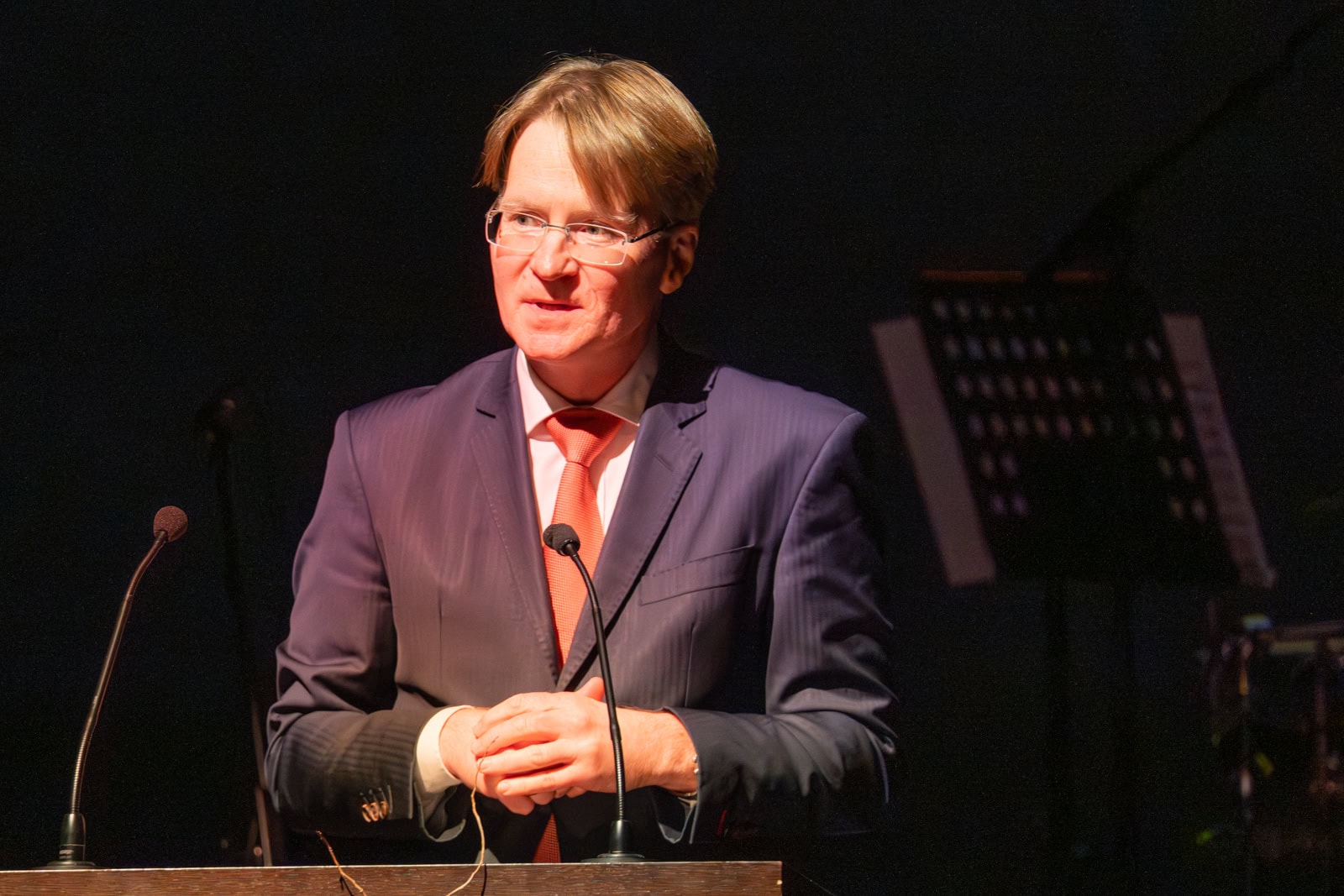 Ein Mann in Anzug, der bei einer Veranstaltung im Rampenlicht spricht.