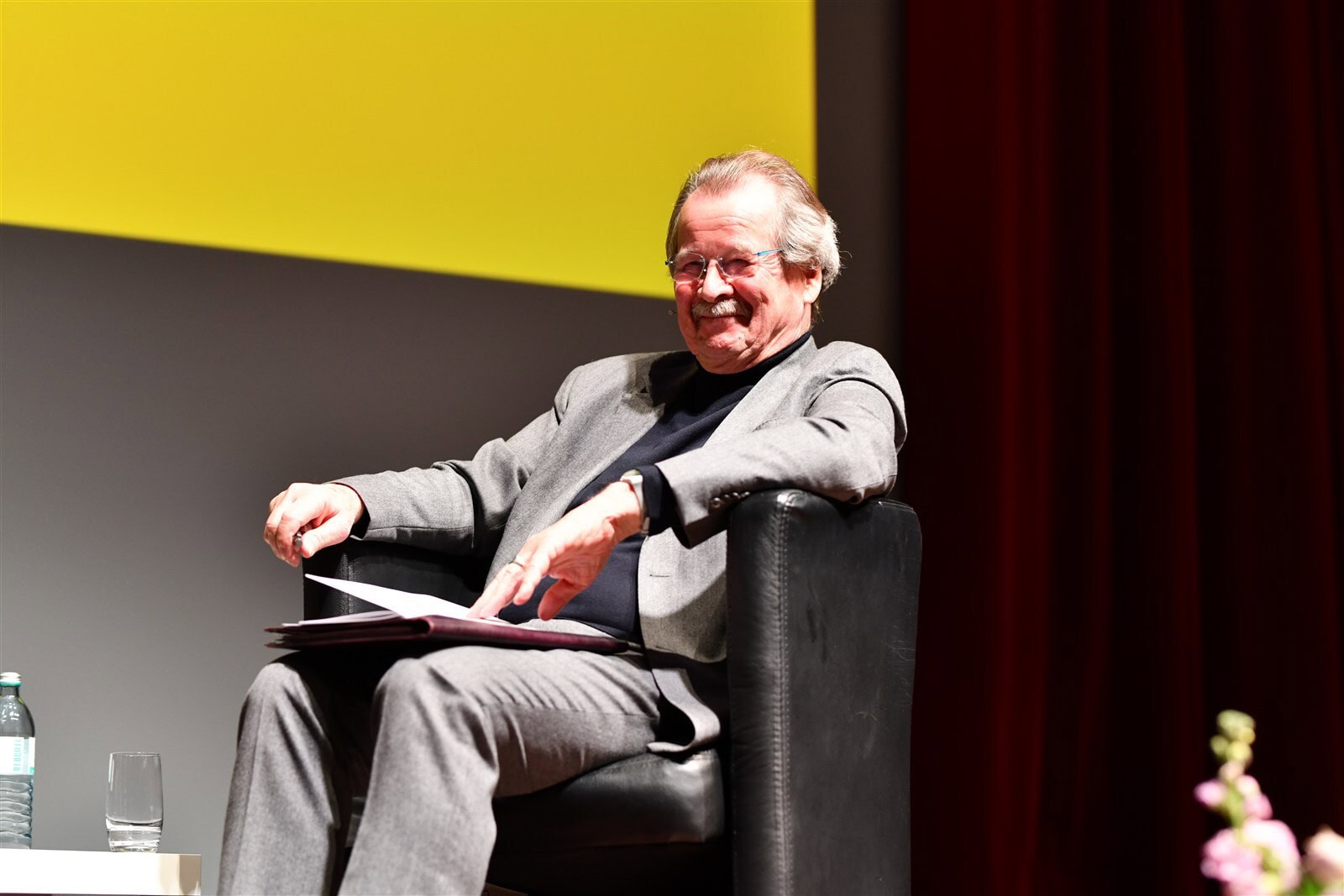 Erfahrener Richter bei Diskussion auf Veranstaltung, sitzt entspannt in einem schwarzen Sessel, im Hintergrund gelbe Wand mit roten Vorhängen, professionelles Ambiente für Rechtsexpertise.