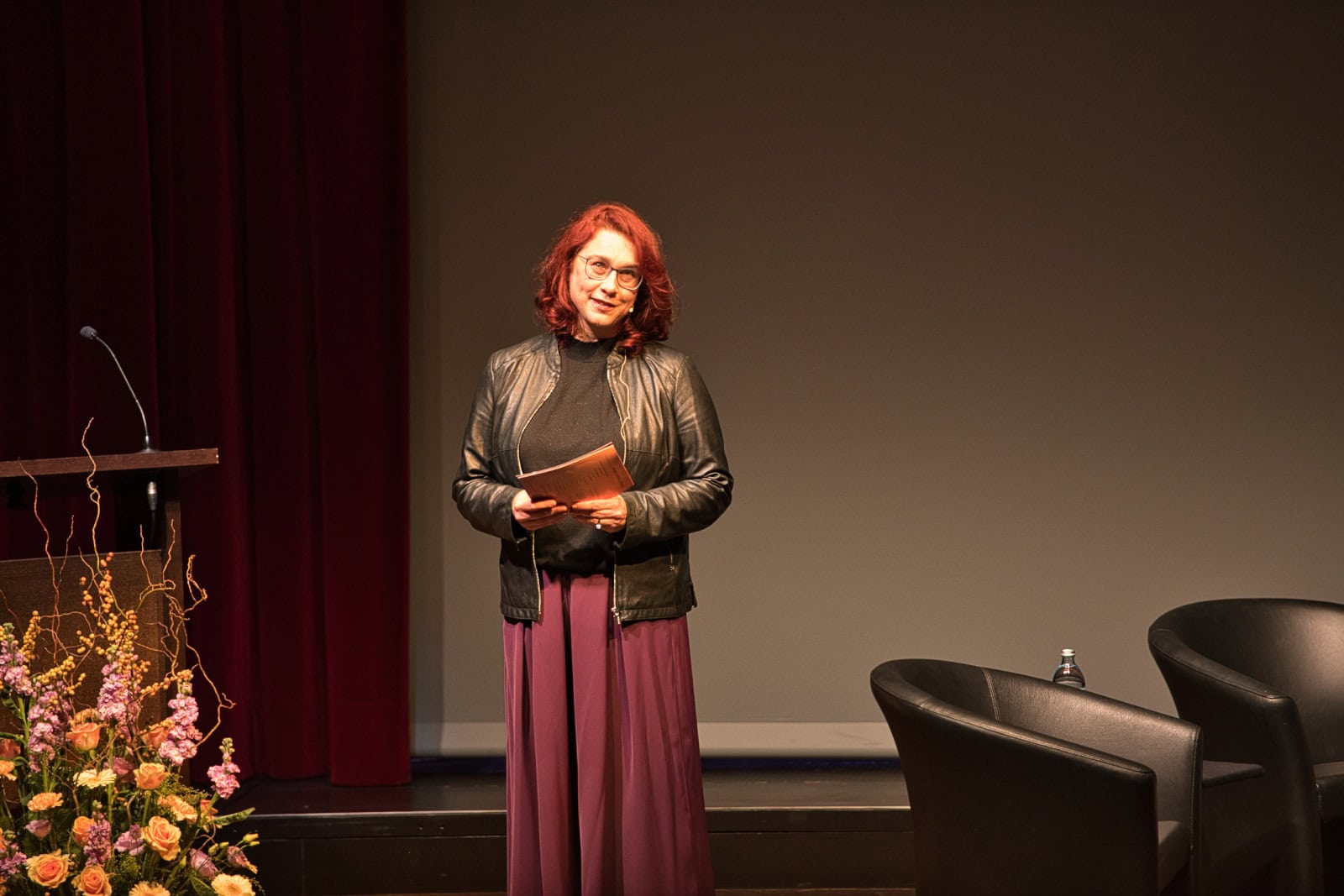 Moderation bei einer Veranstaltung der Richtervereinigung mit einer Frau auf der Bühne, die ein Notizbuch hält, vor einem leeren Hintergrund, professionelle Event-Atmosphäre, Fokus auf Gleichstellung und Berufsstand.