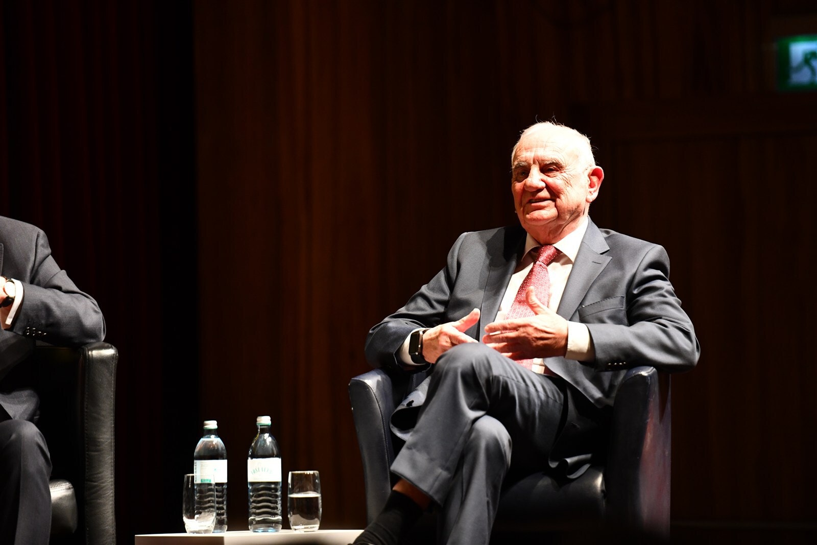 Erfahrener Richter im Gespräch bei Fachveranstaltung, sitzend in schwarzem Anzug, auf einem Panel, hinter ihm eine dunkle Holzvertäfelung, auf dem Tisch Wasserflaschen und Gläser.