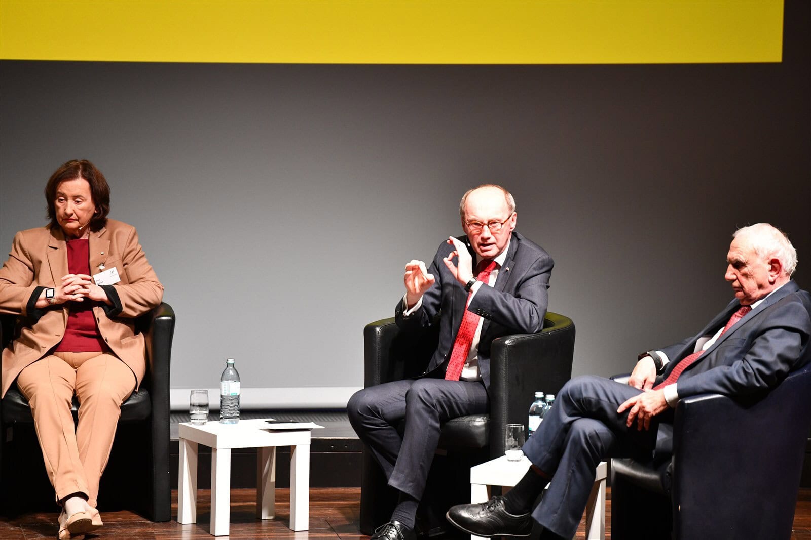 Drei Richter bei Diskussion im Bankett, im Mittelpunkt ein Mann, moderierend, mit Wasserflasche und Becher vor ihnen, professionelles Panel, formelle Veranstaltung, Fokus auf Rechtsprechung.