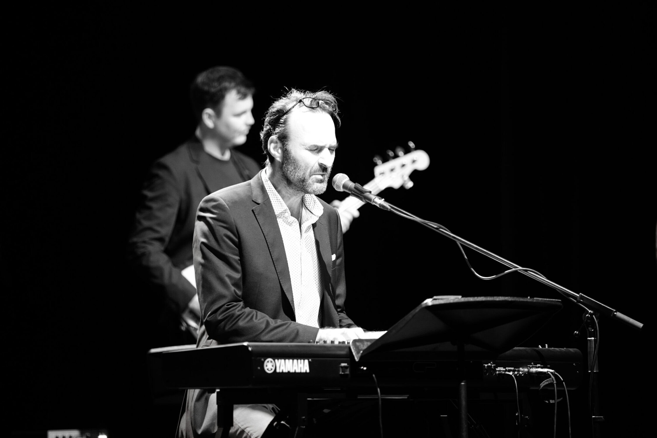 Erfahrener Musiker singt und spielt Keyboard bei einem Konzert, unterstützt von Bassisten im Hintergrund, in einer schwarzen und weißen Fotografie.