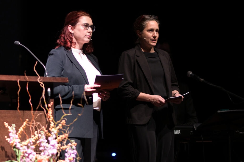 Zwei Frauen bei einer Veranstaltung, die durch die Richtervereinigung präsentiert wird, mit Mikrofonen und Notizen, im dunklen Hintergrund, professionelle Atmosphäre.